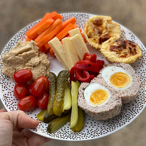 Carrots, gherkins, plum tomatoes, red peppers, cheese sticks, ham and pineapple muffins, scotch eggs and hummus on plate