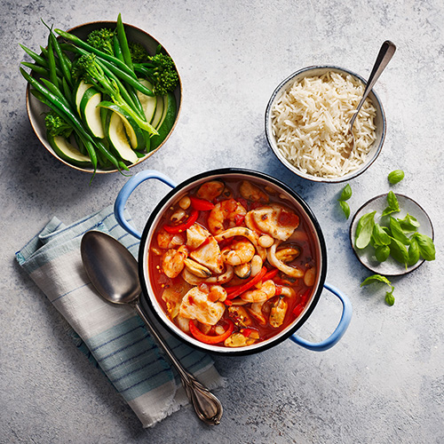 Italian seafood stew with rice and green vegetables in a blue pot on a white background
