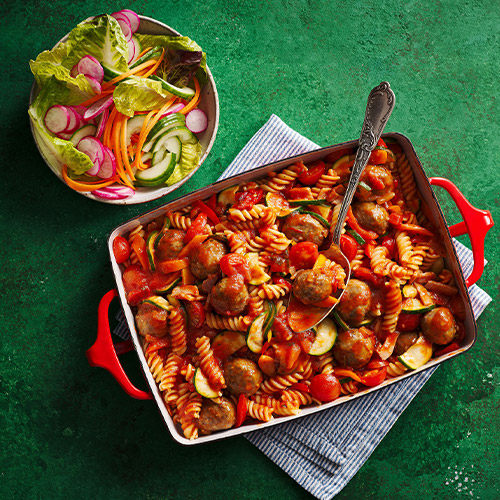 Meatball pasta bake with a side salad in a red roasting pan on a green background