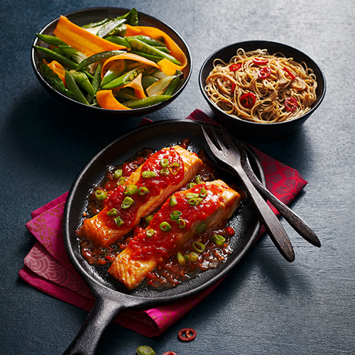 Slimming World sweet chilli salmon in black pan. A side of stir fried veg and noodles in black bowls on a grey background