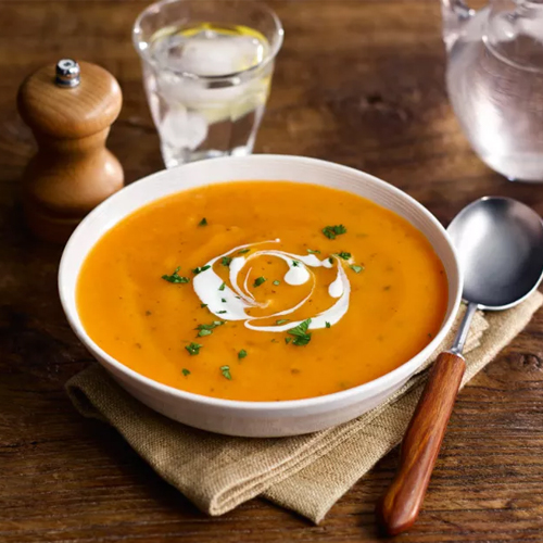 Carrot and coriander Slimming World soup in a white bowl on a brown table with napkin, spoon and a glass of water on the side