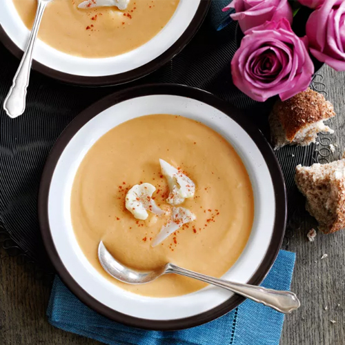 Cauliflower cheese Slimming World soup in black-rimmed bowl with spoon on a wooden table with bread and pink roses