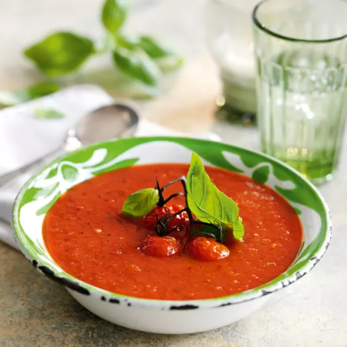 Roasted tomato and basil Slimming World soup in white bowl with green floral pattern on a white tablecloth with a glass of water and a silver spoon