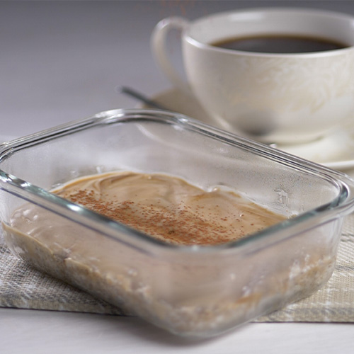 Tiramisu Weetabix cheesecake in a glass container on a grey worktop. White mug filled with black coffee in the background.