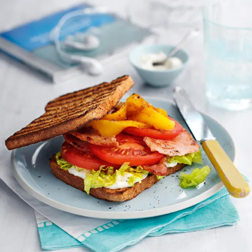 Slimming World Bacon, lettuce and tomato sandwich. The sandwich consists of two slices of toasted wholemeal bread, topped with lettuce, tomato slices, bacon and yellow peppers. It sits on a light blue plate resting on a white table.