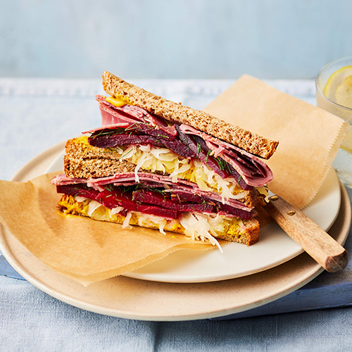 Slimming World New York Deli style corned beef and mustard sandwich.  The sandwich consists of two slices of whole wheat bread filled with beef, sauerkraut and marinated ham.  It sits on a light brown baking sheet on top of a white plate.  The background is light gray.