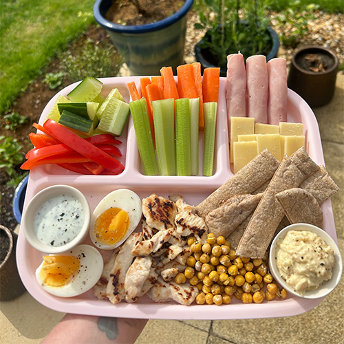 A pink picnic platter filled with sliced red peppers, cucumber, carrots, celery, ham and cheese slices, boiled eggs, roasted chicken, chickpeas, pitta bread, hummus and tzatziki