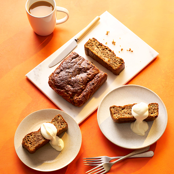 Banana bread loaf on a chopping board, with two slices shown served with fat-free natural Greek yogurt and a cup of tea
